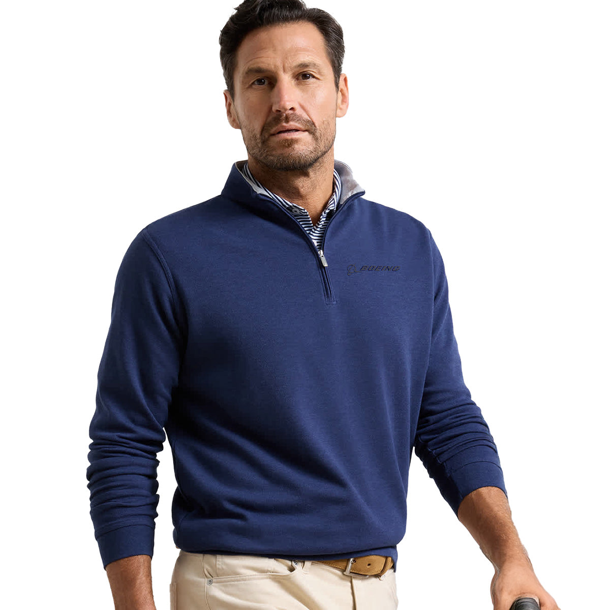A man with short brown hair and light stubble wears a blue Peter Millar Boeing Men’s Crown Comfort Knit Quarter-Zip over a striped collared shirt and holds a golf club. He stands against a plain white background, looking slightly to the side.