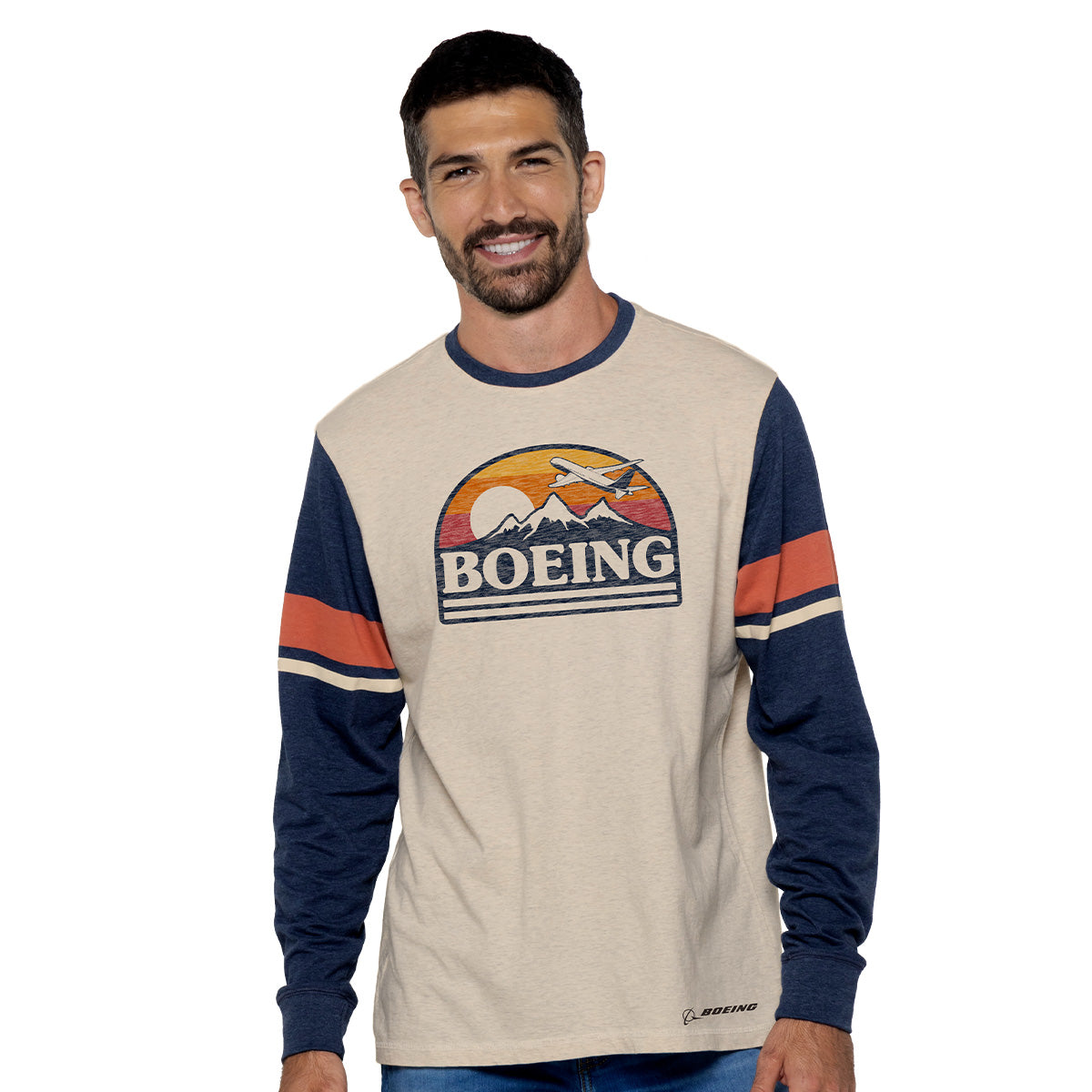 A man confidently smiles in a beige and navy Boeing Horizon Mens Long Sleeve T-Shirt by Boeing, featuring arm stripes, a mountain and plane design, and Boeing Horizon displayed at the center, reflecting his passion for aviation.