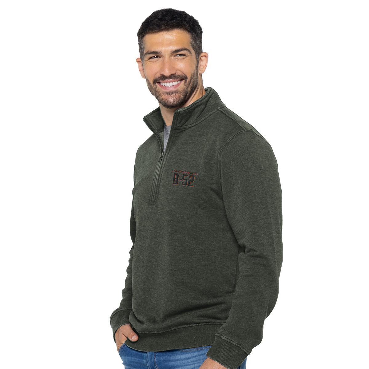 A man with a beard smiles in a dark green Boeing B-52 Mens Quarter-Zip sweater made from cozy cotton/poly fleece, featuring B-52 print. He stands against a white backdrop with his hands in blue jean pockets.