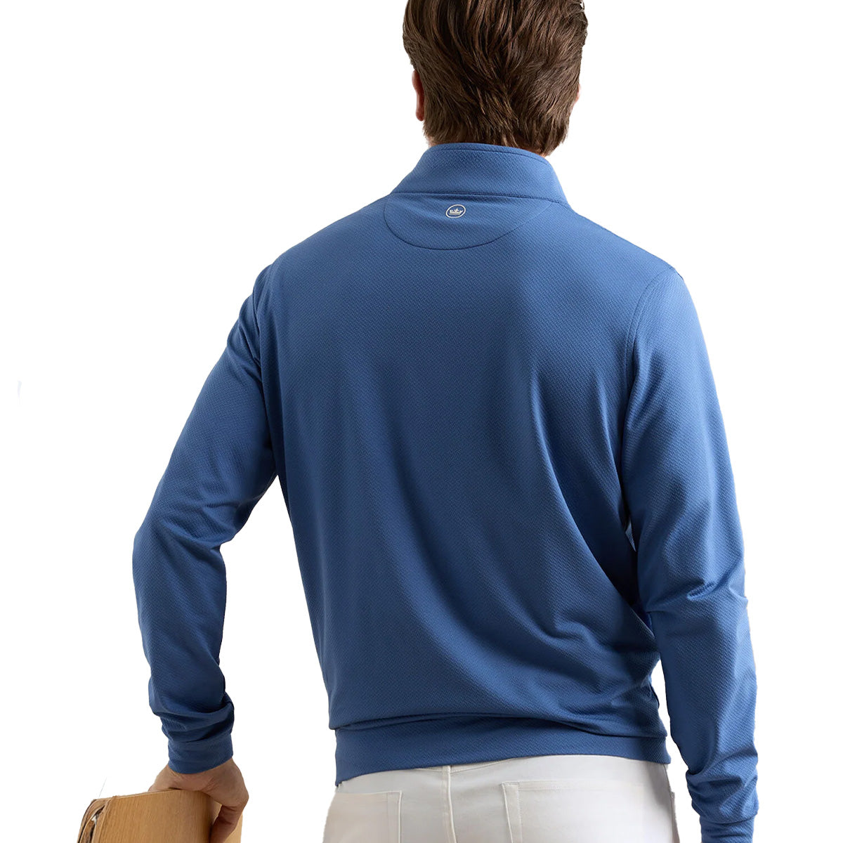 A person wearing a blue Boeing Peter Millar Men’s Perth Cross-Hatch Quarter-Zip and white pants is seen from behind, holding a tan bag in their left hand against a plain white background.