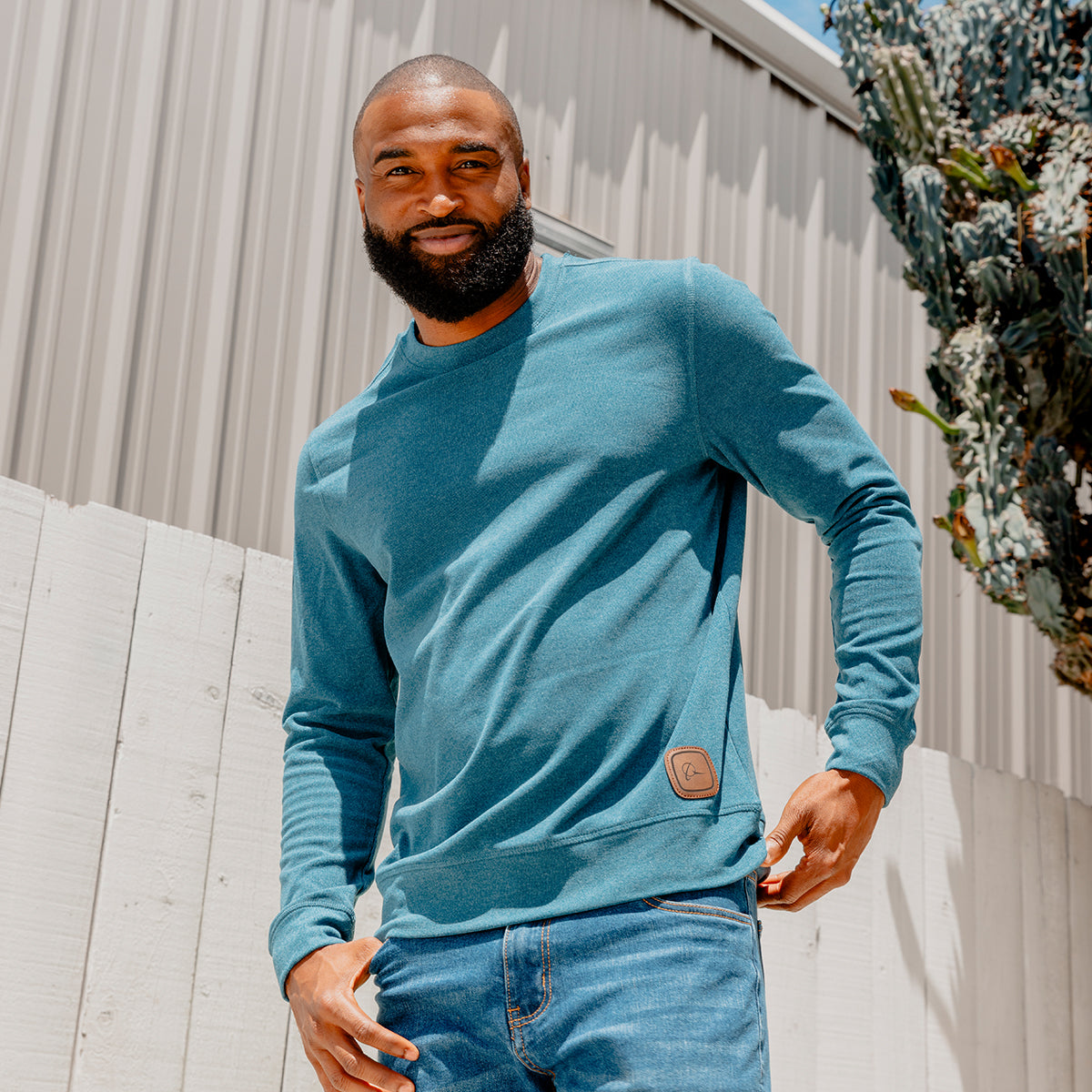 A bearded man stands outdoors by a white fence and a metal building, with a cactus in the background, wearing the Boeing Symbol Patch Unisex Crewneck Sweatshirt from Boeing along with jeans.