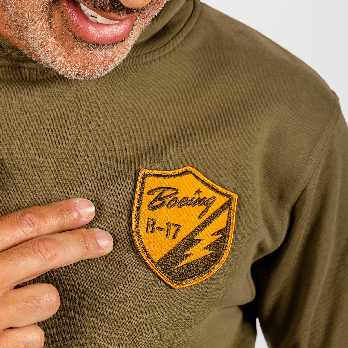 A person points to a yellow patch on an olive green BOEING Heritage B-17 Flying Fortress Unisex Hoodie, featuring aviation-inspired apparel. The patch displays “Boeing B-17” with a star and lightning bolt design, while only the lower face and hand are visible.