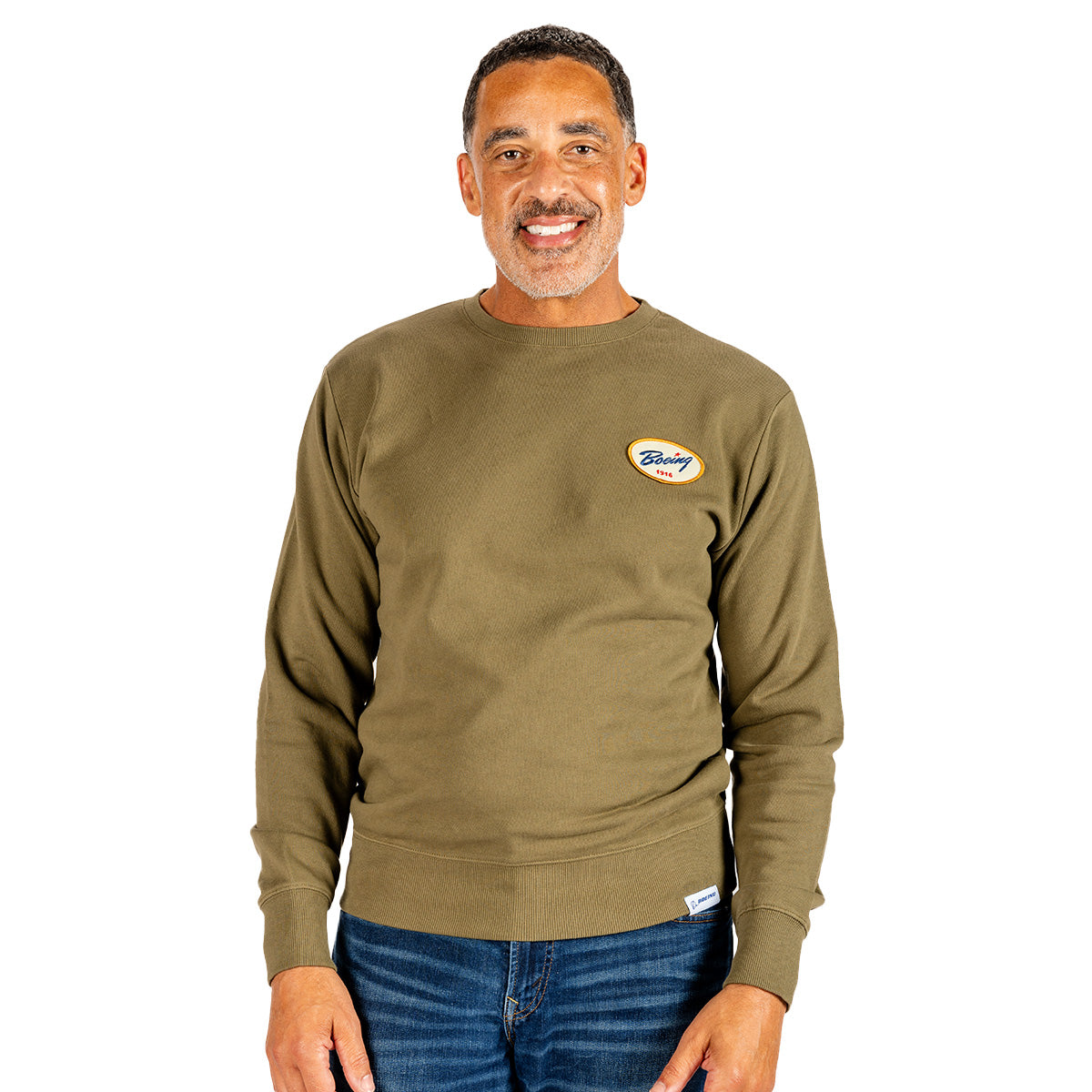 A man in a greenish-brown Boeing Heritage Airplane Company Unisex Crewneck Sweatshirt and blue jeans smiles while standing against a white background.