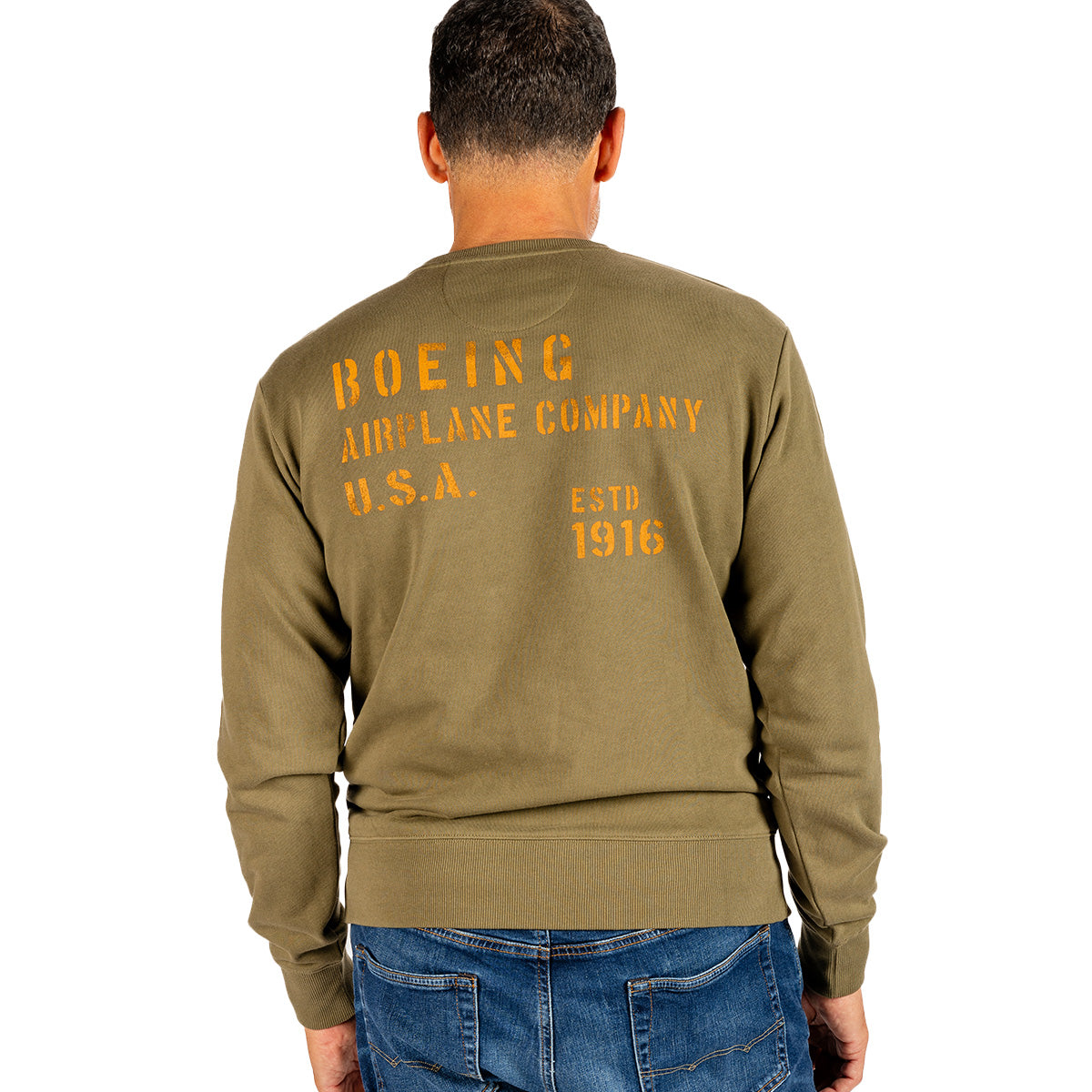 A man faces away, wearing an olive green Boeing Heritage Airplane Company Unisex Crewneck Sweatshirt in French terry with yellow heritage script applique, paired with blue jeans against a white background.
