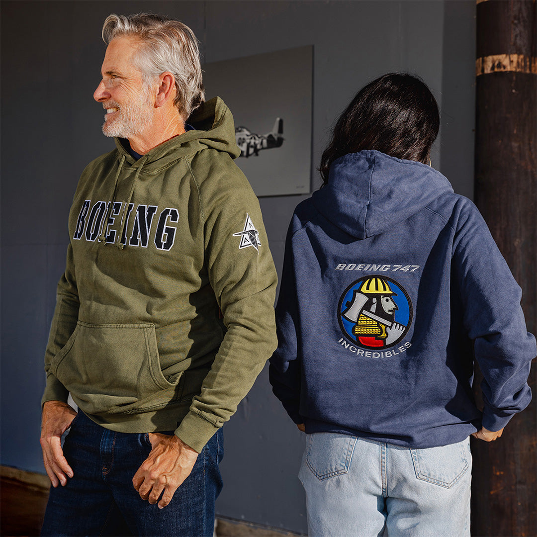 Man and woman lifestyle wearing Boeing heritage hoodies. The man is wearing a green P-51 hoodie with large Boeing logo embroidered across the chest and the woman is wearing a blue 747 hoodie with the Incredibles embroidered patch on the back.