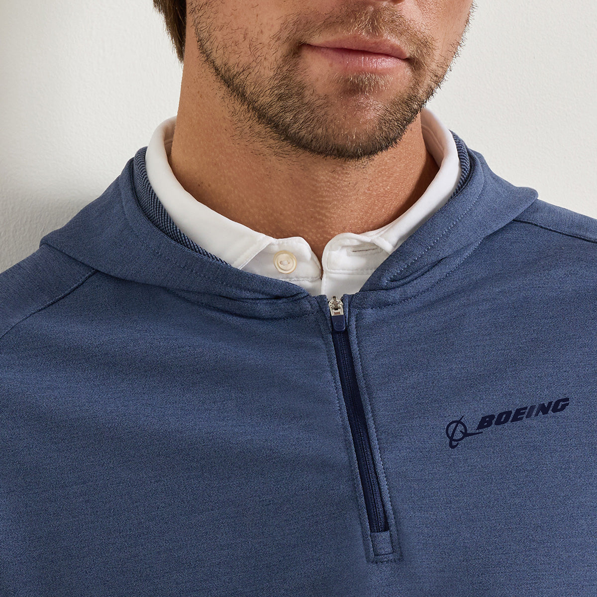 A man is seen wearing the Peter Millar Boeing Men’s Pine Quarter-Zip Performance Hoodie in blue, layered over a white collared shirt. The hoodie features the BOEING logo on the left chest and is designed to provide comfort and flexibility for everyday wear. Only the lower half of his face and upper torso are visible.