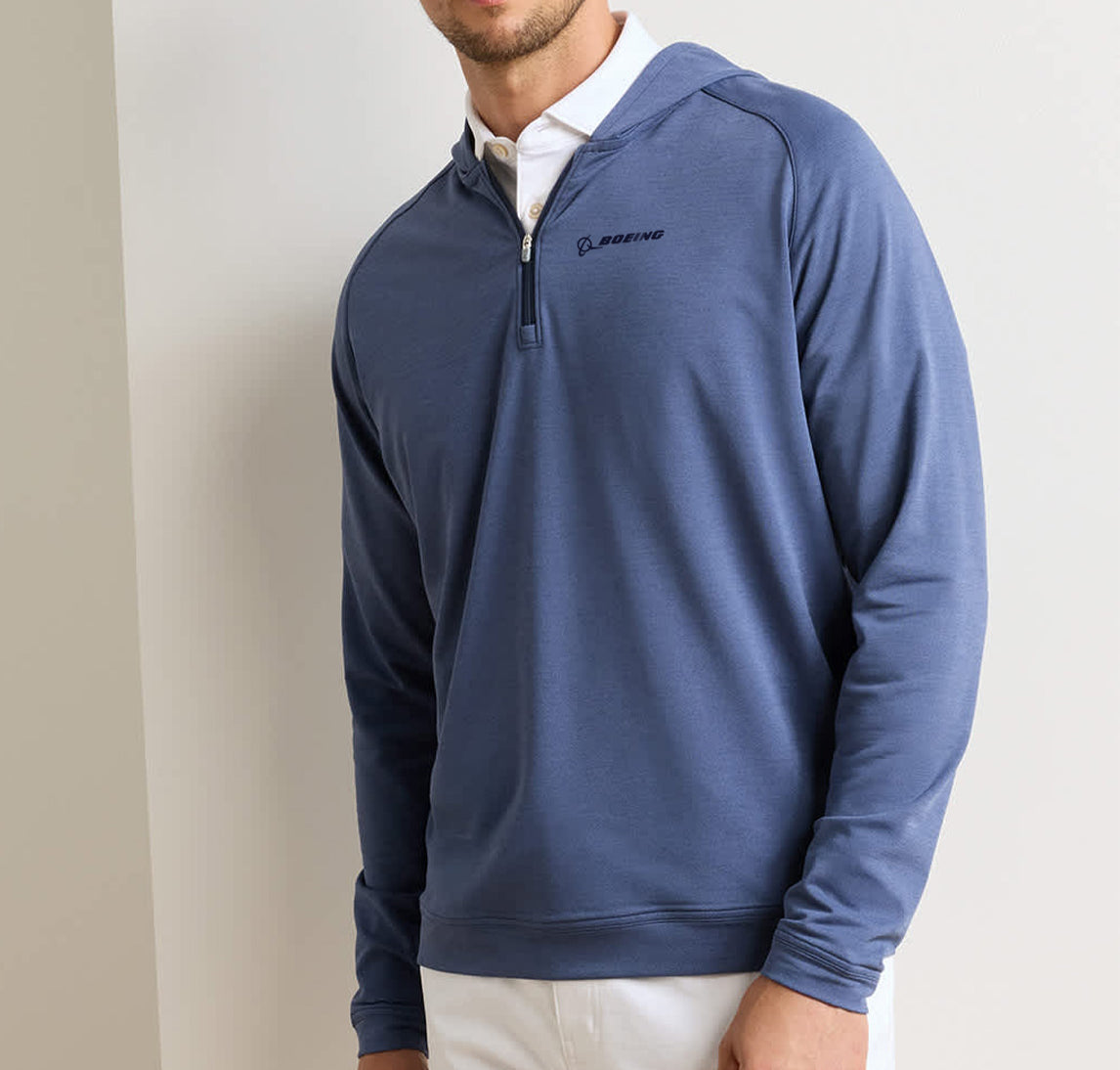 A man wearing the Peter Millar Boeing Men’s Pine Quarter-Zip Performance Hoodie—a blue, four-way stretch pullover with Boeing branding—over a white collared shirt and white pants stands against a light-colored wall.