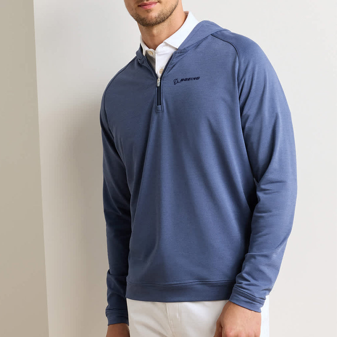 A man wearing the Peter Millar Boeing Men’s Pine Quarter-Zip Performance Hoodie—a blue, four-way stretch pullover with Boeing branding—over a white collared shirt and white pants stands against a light-colored wall.