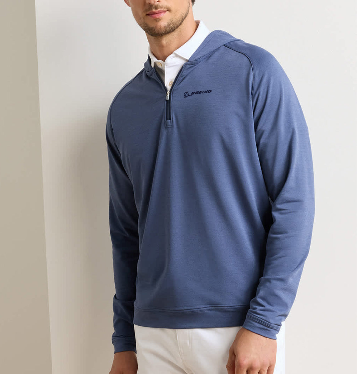 A man wearing the Peter Millar Boeing Men’s Pine Quarter-Zip Performance Hoodie—a blue, four-way stretch pullover with Boeing branding—over a white collared shirt and white pants stands against a light-colored wall.