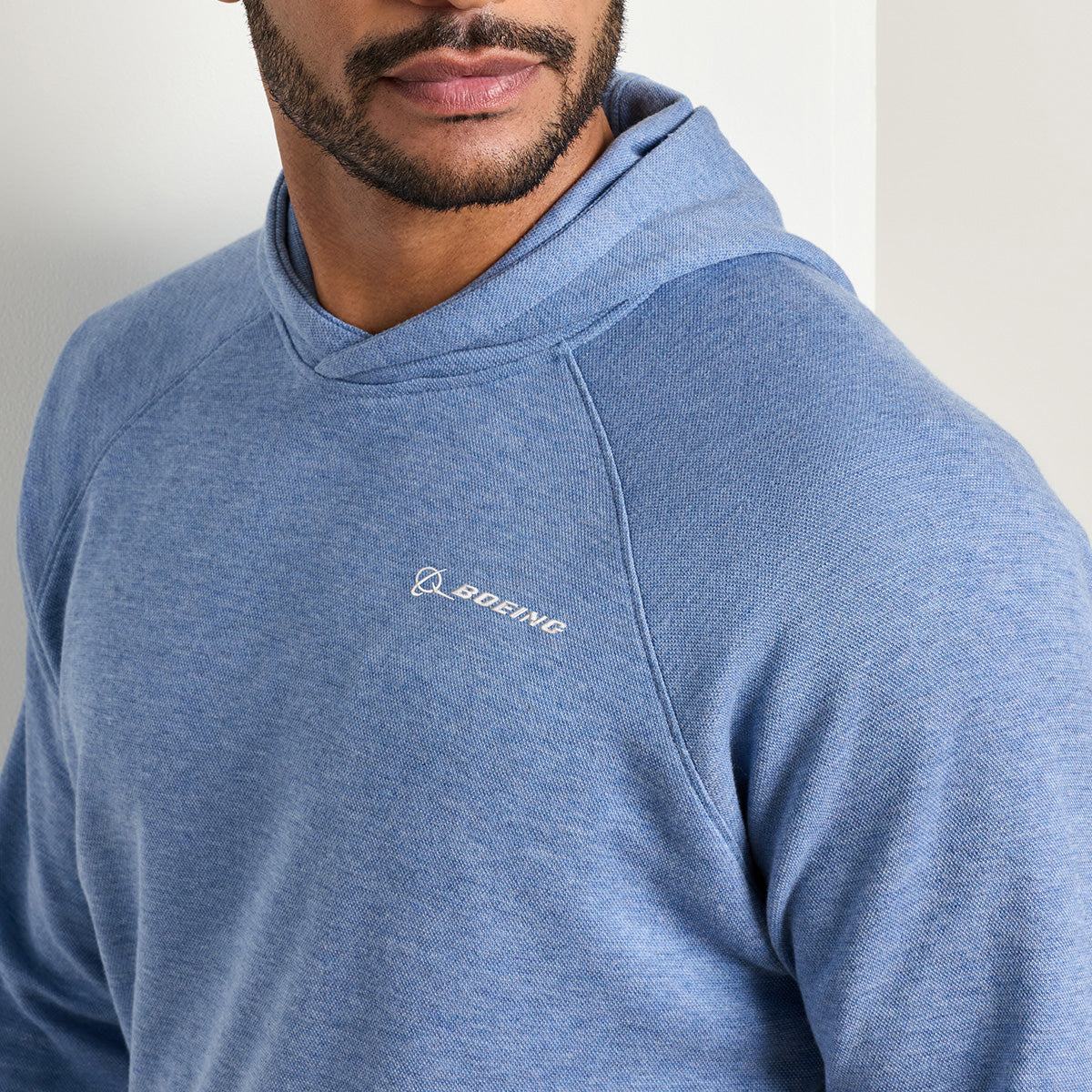 A man wearing a Peter Millar Boeing Men’s Crown Comfort Knit Hoodie in light blue, featuring the BOEING logo on the upper chest, stands against a white wall.