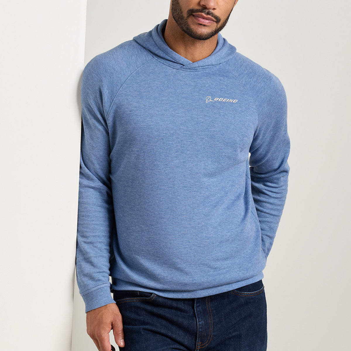 A man wearing the Peter Millar Boeing Men’s Crown Comfort Knit Hoodie by BOEING, featuring the Boeing logo on the chest, stands against a light-colored wall with one hand in his pocket.