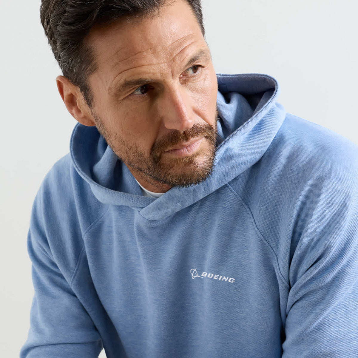 A man with brown hair and a beard wears the Peter Millar Boeing Men’s Crown Comfort Knit Hoodie in light blue, featuring the Boeing logo on the chest. He gazes slightly to his right against a plain background.