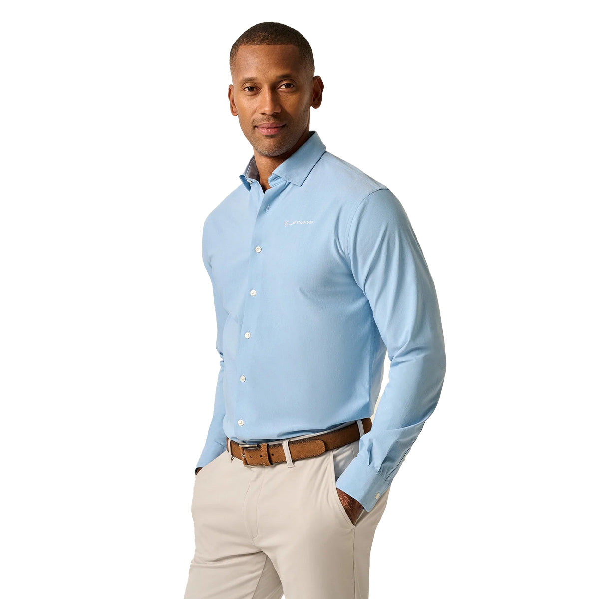 A man stands with his hands in his pockets, wearing a wrinkle-resistant BOEING johnnie-O Men’s Travis Performance Button Down Shirt in light blue, beige pants, and a brown belt, looking at the camera against a plain white background.