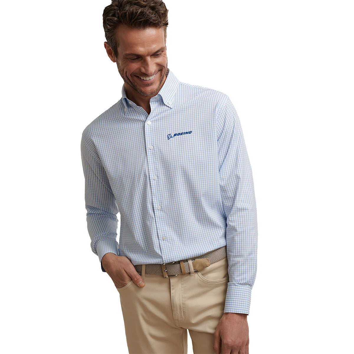 A man wearing a Peter Millar Boeing Men’s Hanford Twill Sport Shirt with a blue and white check pattern, beige pants, and a woven belt smiles while looking down, one hand in his pocket. Brand: BOEING.