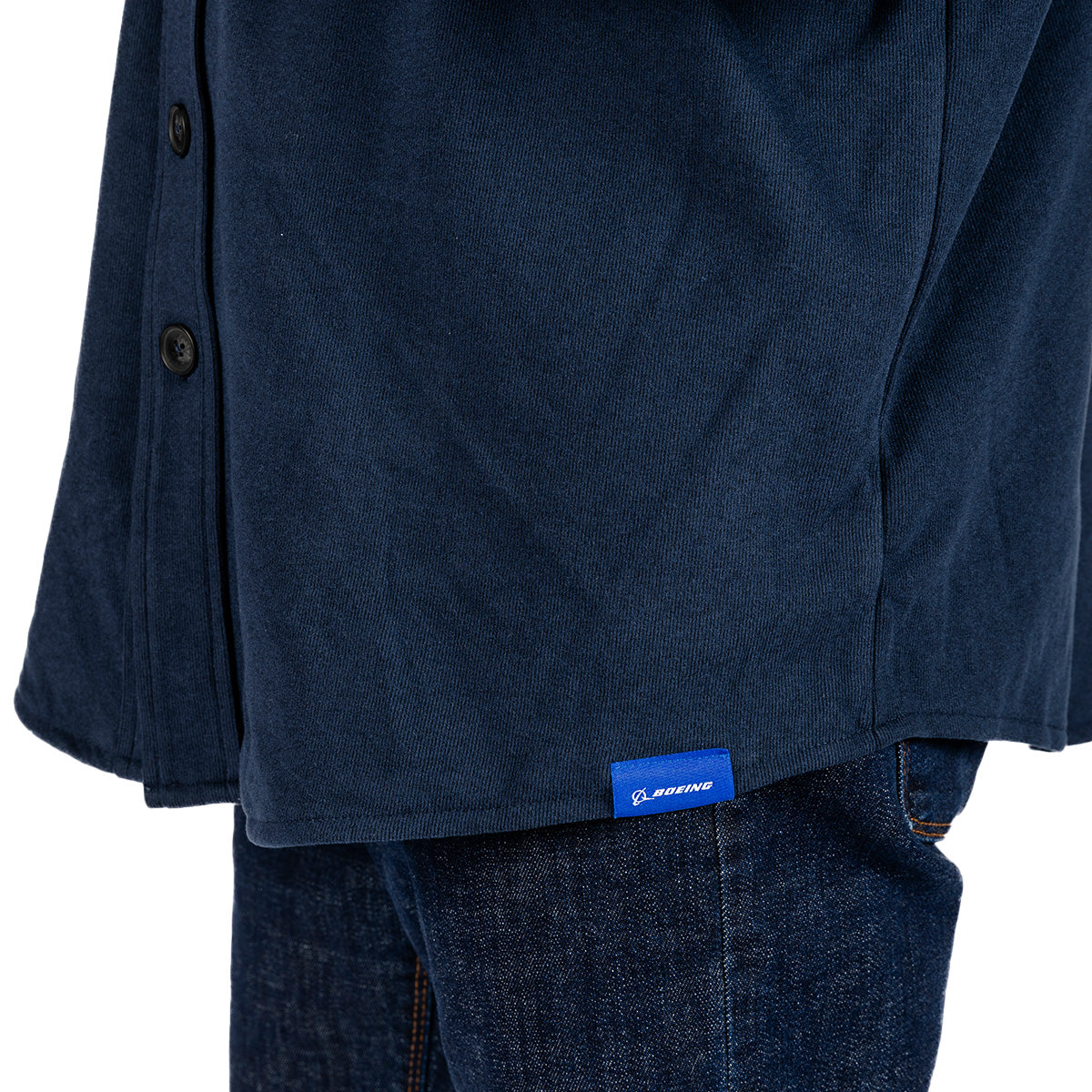 A close-up of a person wearing the Boeing Unisex Journey Overshirt in navy blue spandex blend with black buttons, paired with blue jeans. A small blue tag featuring the BOEING logo is visible at the bottom of the overshirt.