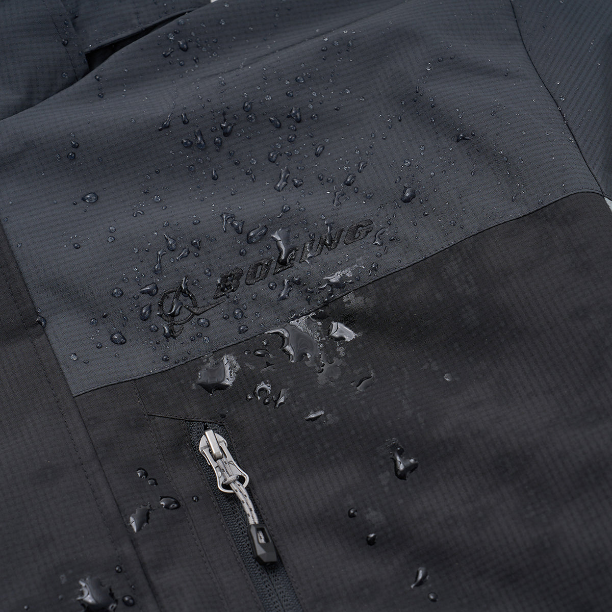 Close-up of the Boeing Men's Trifecta Ripstop Jacket by BOEING, showing water droplets on its water-resistant surface. A partially visible zipper and subtle BOEING logo emphasize the jacket's durable, high-quality construction.