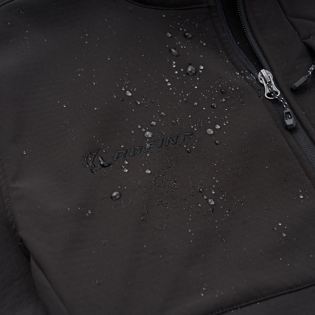 Close-up of a Boeing Men's Softshell Jacket in black, featuring water-resistant material with droplets on the surface. The BOEING logo is embroidered near the center, and a zipper is partially visible on the right.