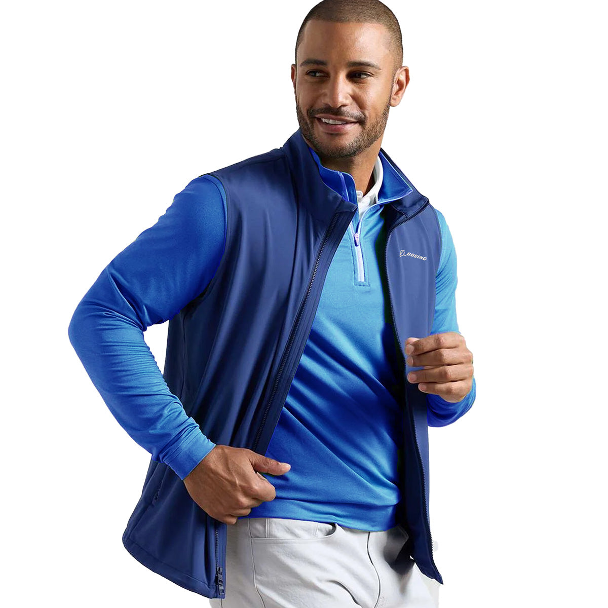 A man in a blue long-sleeve shirt, light pants, and a BOEING Peter Millar Men’s Dunes Performance Vest smiles as he unzips the windproof, water-resistant vest. He has a short haircut and trimmed beard against a plain white background.
