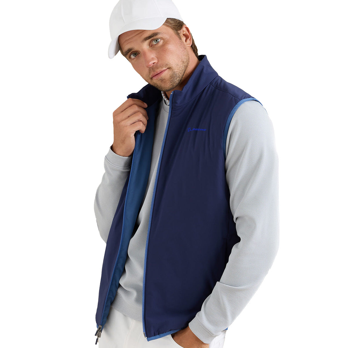 A man wearing a white cap, light gray long-sleeve shirt, and a navy blue Peter Millar Boeing Men’s Ridge Vest by BOEING poses holding the vest collar and looks at the camera against a white background.