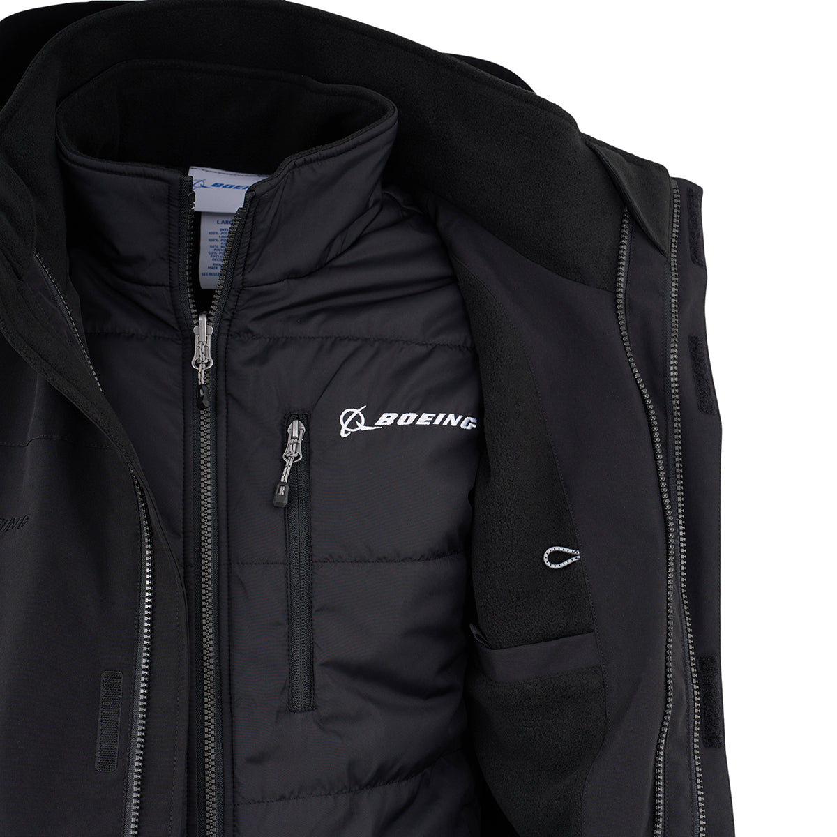 A close-up of the Boeing Men's 3-in-1 Systems Jacket by BOEING shows a partially unzipped black outer layer, revealing a zippered inner layer with the white embroidered Boeing logo on the left chest and detailed inner pockets.