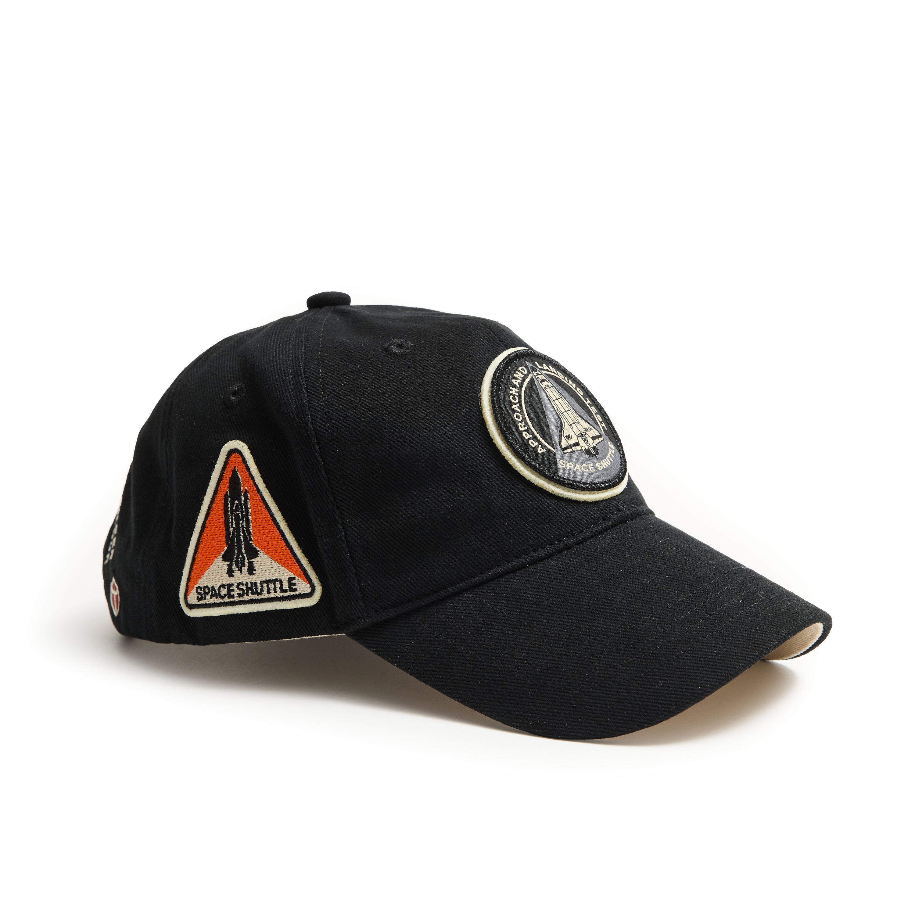 The Boeing Red Canoe NASA ALT Shuttle Hat is a black baseball cap with two embroidered patches: a triangular SPACE SHUTTLE patch and a circular patch celebrating a space mission, making it perfect for space enthusiasts.