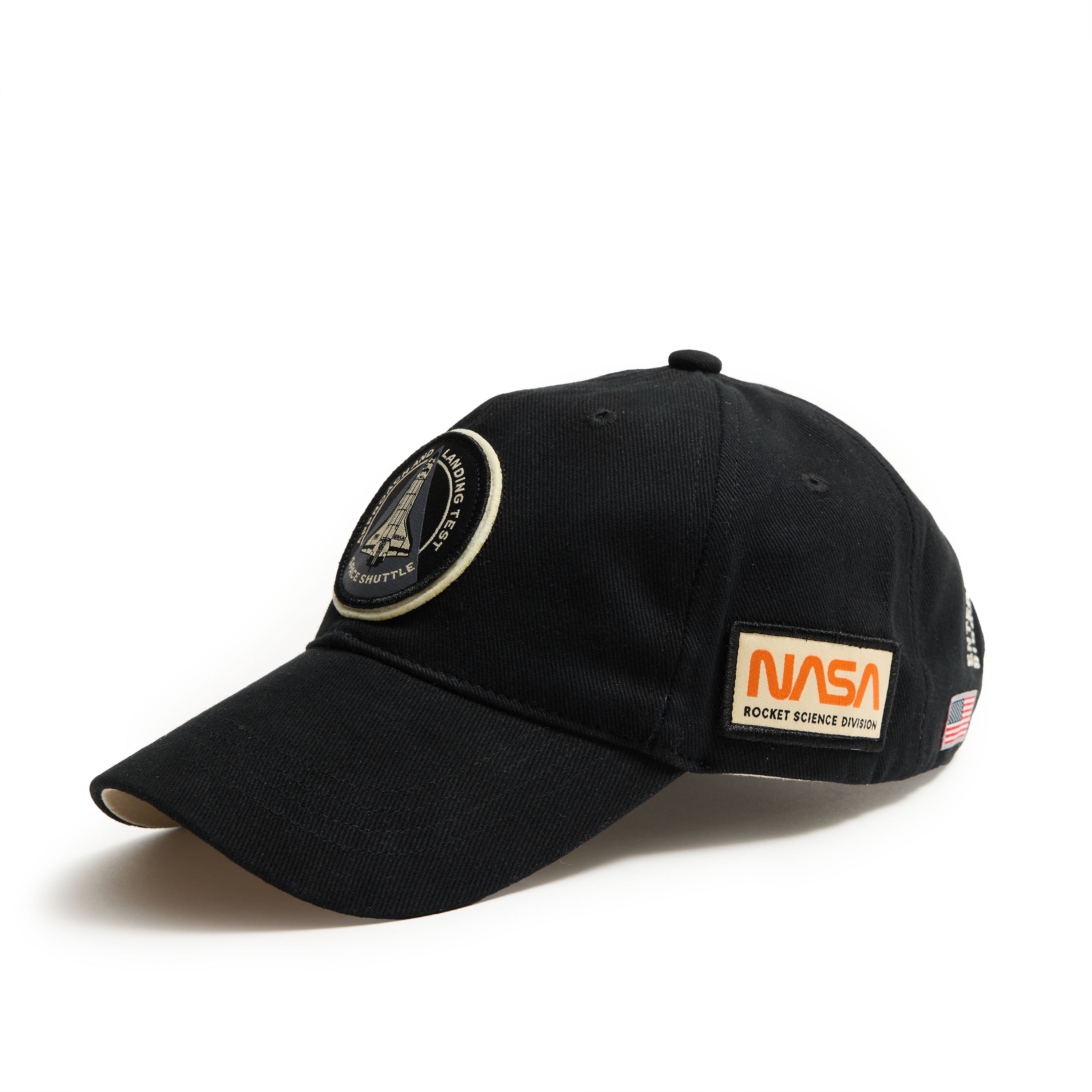 This Boeing Red Canoe NASA ALT Shuttle Hat is a black cap adorned with embroidered NASA patches, including a shuttle and ROCKET SCIENCE DIVISION text, plus an American flag patch on the side. Perfect for space enthusiasts, its artfully angled on a white background.