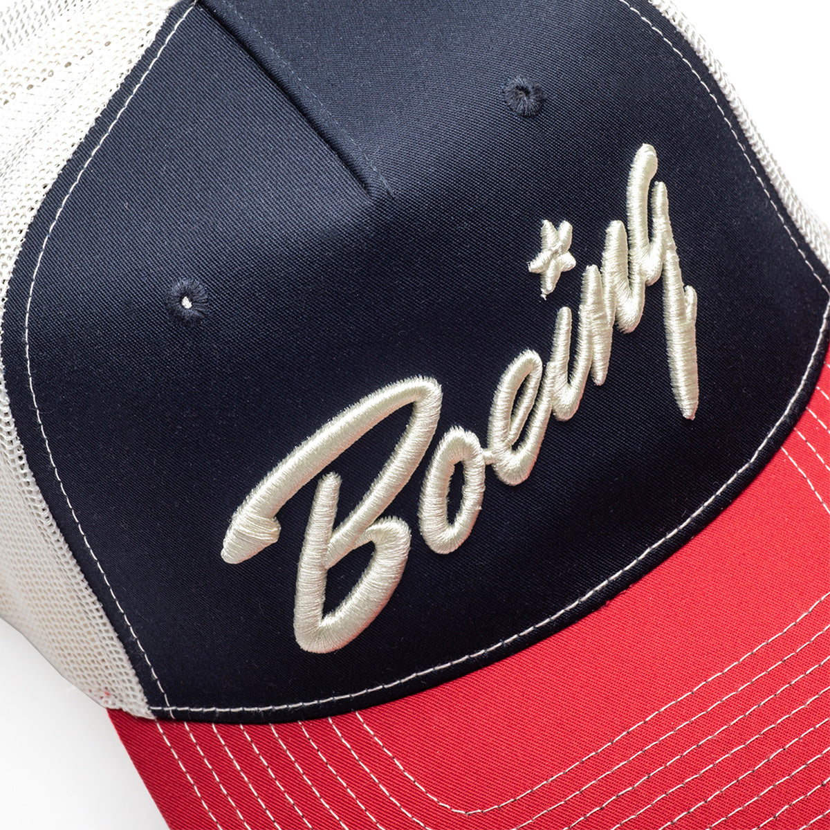 A close-up of the Richardson Boeing Script Logo Trucker Hat by BOEING, featuring a navy front, white mesh sides, red bill with white stitching, the embroidered Boeing Script logo in white, and a Softsnap Adjustable Backstrap.