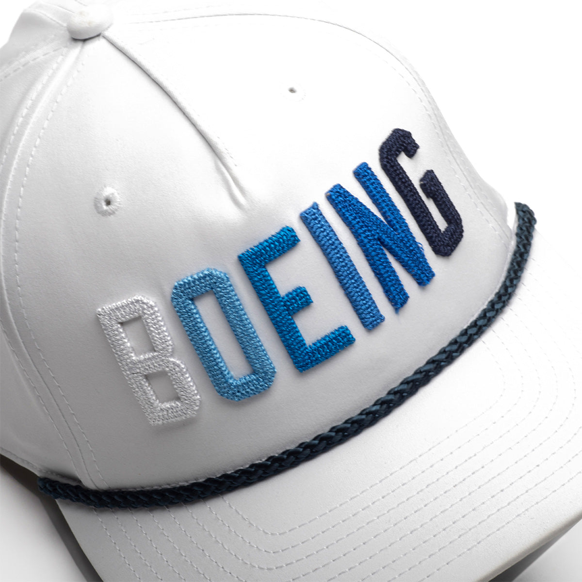 The Richardson Boeing Golf Hat by BOEING is a white performance polyester cap with "BOEING" boldly embroidered in blue and white, plus a dark braided rope across the visor for a sporty look.