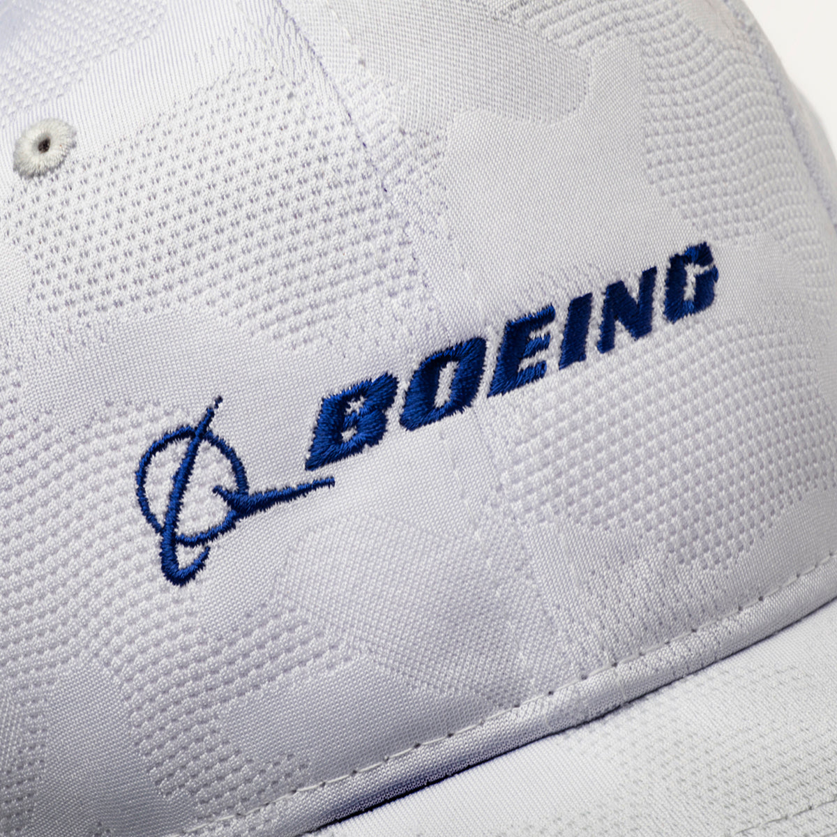The Boeing Signature Logo Camo Hat by BOEING showcases a white design with blue embroidered text, subtle camo pattern fabric, visible stitching, and a snapback closure for adjustable fit.