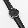 The Boeing Silver Rotating Airplane Watch by Boeing features a minimalist black dial encased in a silver case with a sleek black leather strap. Silver hour markers and hands move elegantly, showcasing timeless style as it rests gracefully on a white surface.