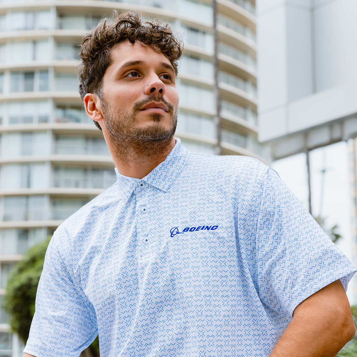 A man with short curly hair and light facial hair wears the Boeing Men's Signature Logo Pro Crossroads Polo by BOEING in light blue, standing outdoors in front of modern buildings and trees.