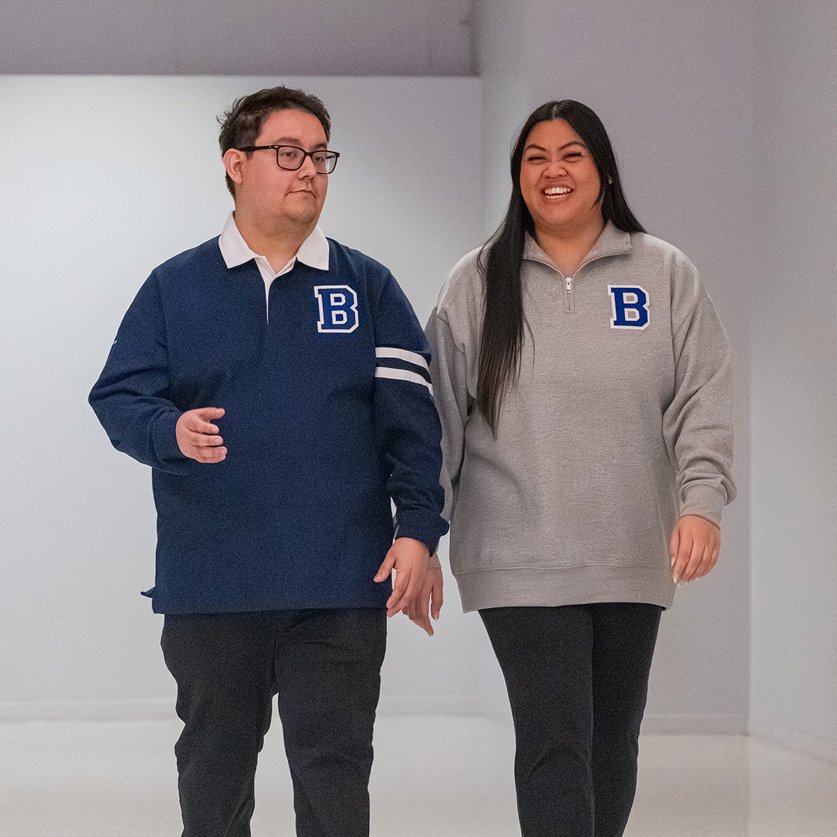 Man and woman wearing Boeing branded Varsity sweatshirts. The man is wearing a navy blue long sleeve polo with white collar and large tonal 'B' patch on the chest and the woman is wearing a grey quarter-zip with large contrast 'B' patch on left chest.