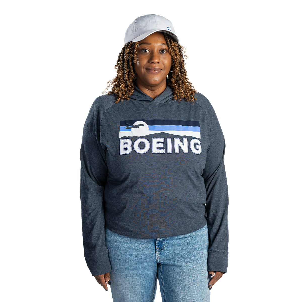 A woman in the Boeing Unisex Boulevard Hoodie by BOEING, paired with light blue jeans and a gray cap, stands against a white background, smiling slightly—showcasing quality basics.