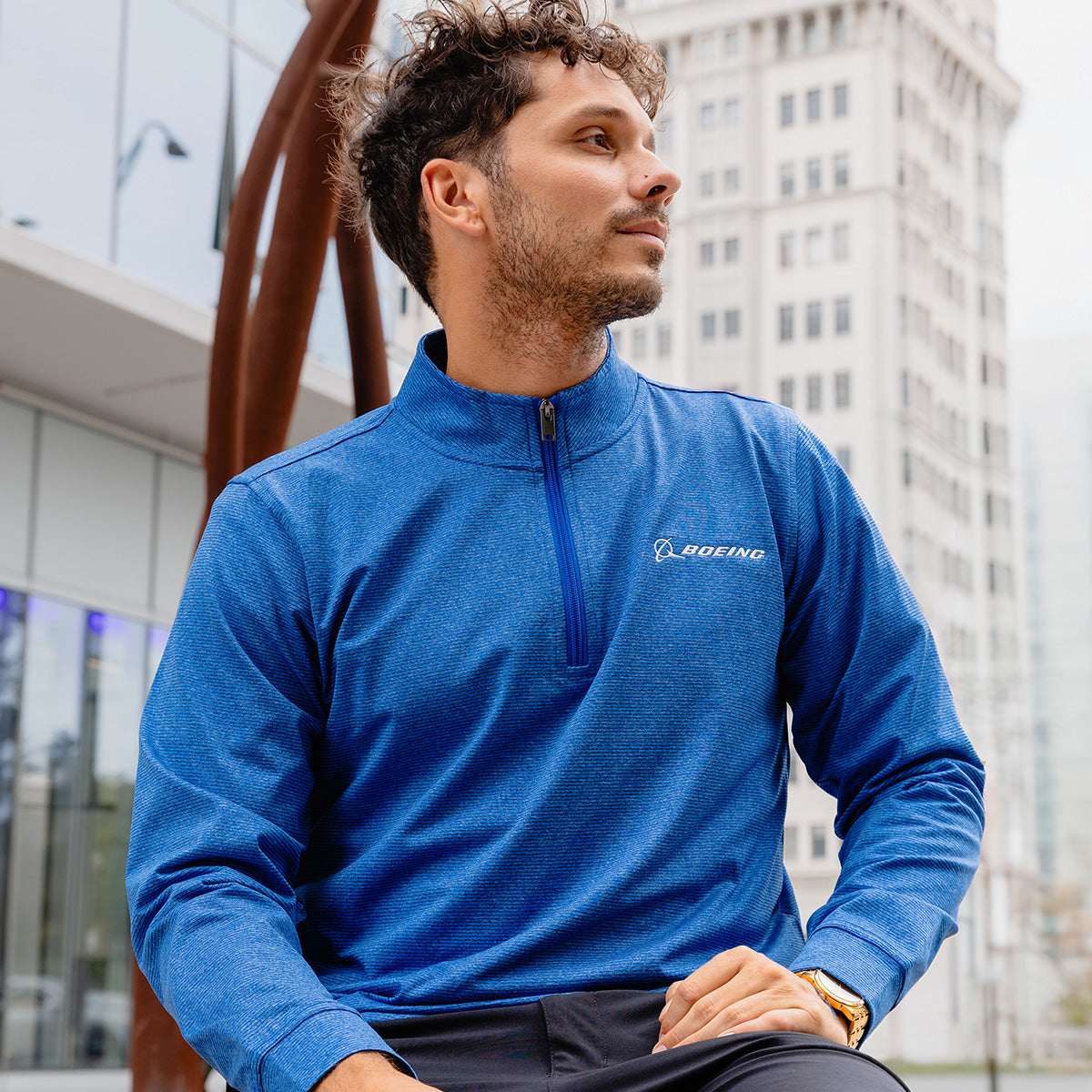 A man wearing the Boeing Men's Signature Logo Stripe Quarter-Zip Pullover by BOEING sits outdoors in a city, modern buildings and sculpture behind him, looking to the side with a thoughtful expression.