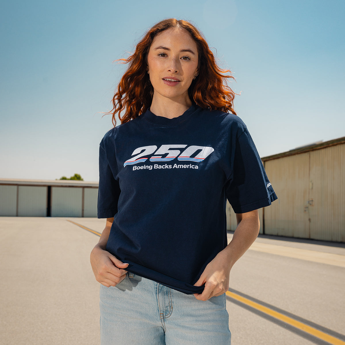 A woman with long, wavy red hair smiles in the sun, wearing a limited-edition Boeing America 250 Unisex T-Shirt by BOEING made from USA cotton. The navy shirt pairs with her light blue jeans as she stands outside.