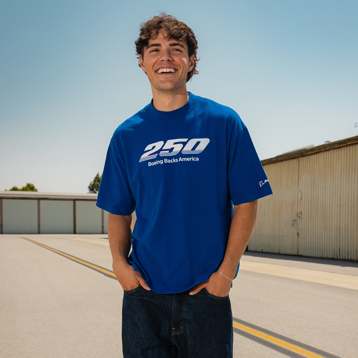 A young man smiles outdoors in a bright blue BOEING America 250 Unisex T-Shirt, crafted from USA cotton and featuring the "250 Bowing Backs America" design. He stands with hands in pockets on a road near industrial buildings.