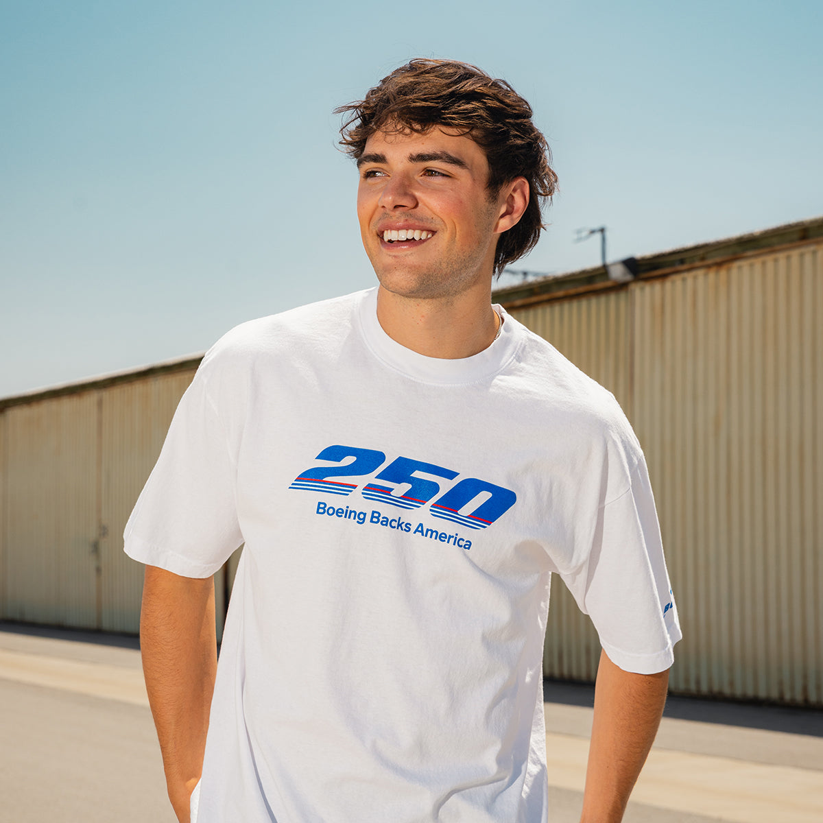 A young man smiles outdoors in a BOEING America 250 Unisex T-Shirt, featuring blue “250 Boeing Backs America” text on USA cotton. The Boeing logo and a corrugated metal building are visible under a clear sky.