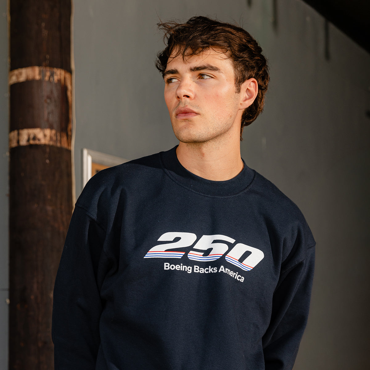 A young man with short brown hair wears a BOEING America 250 Unisex Crewneck featuring bold white and red lettering. He stands against a gray wall, gazing to the side with a serious expression.