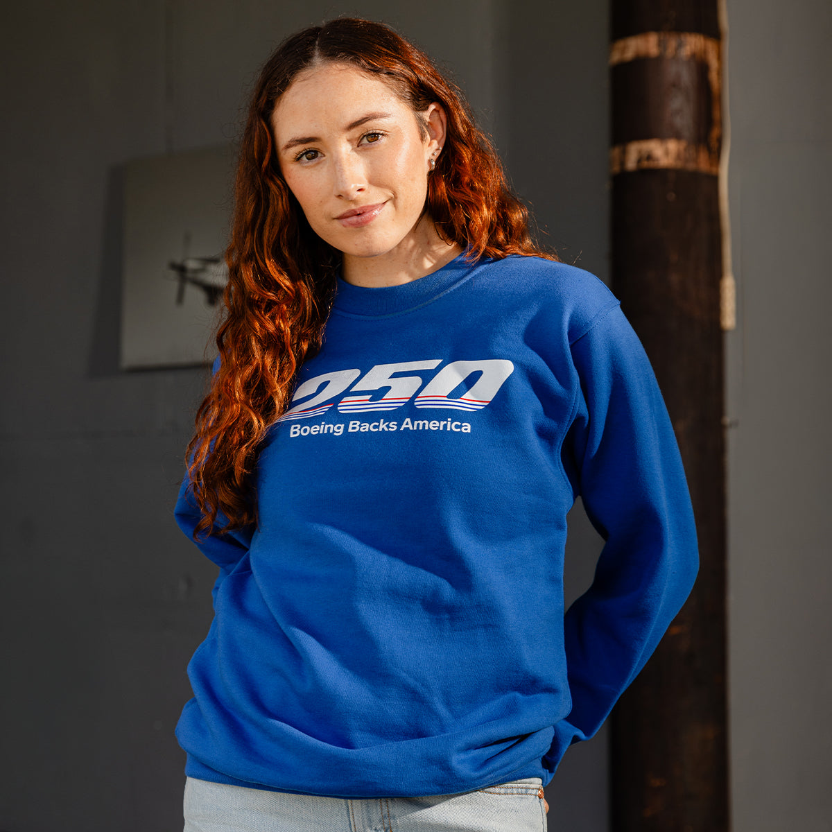 A woman with long red hair wears a BOEING America 250 Unisex Crewneck featuring "250 Boeing Backs America" on the front, posed against a dark background and looking at the camera with a slight smile.