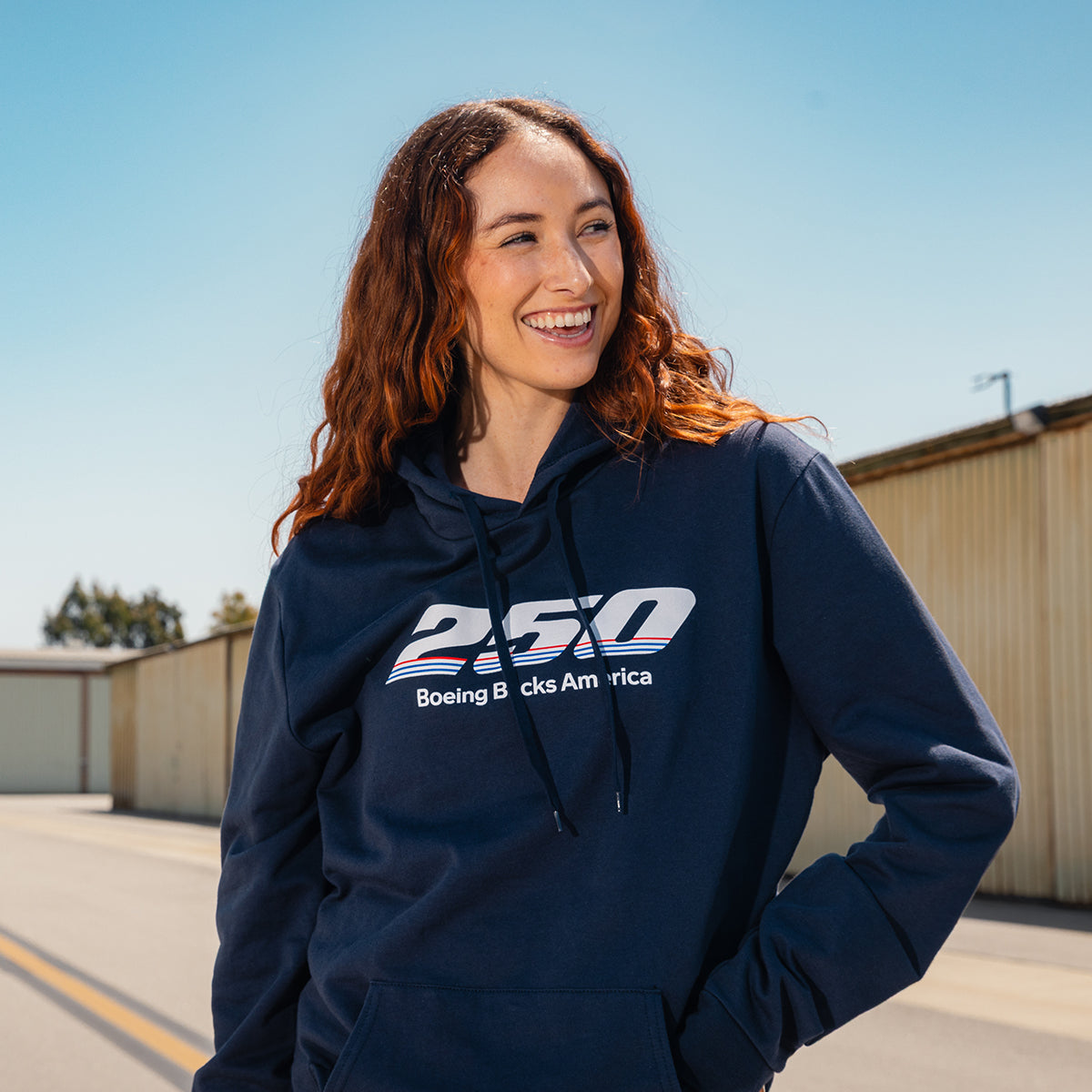 A smiling woman with long red hair wears a BOEING America 250 Unisex Hoodie, made of 100% ring-spun cotton, as she stands outdoors in front of a light-colored industrial building on a sunny day.