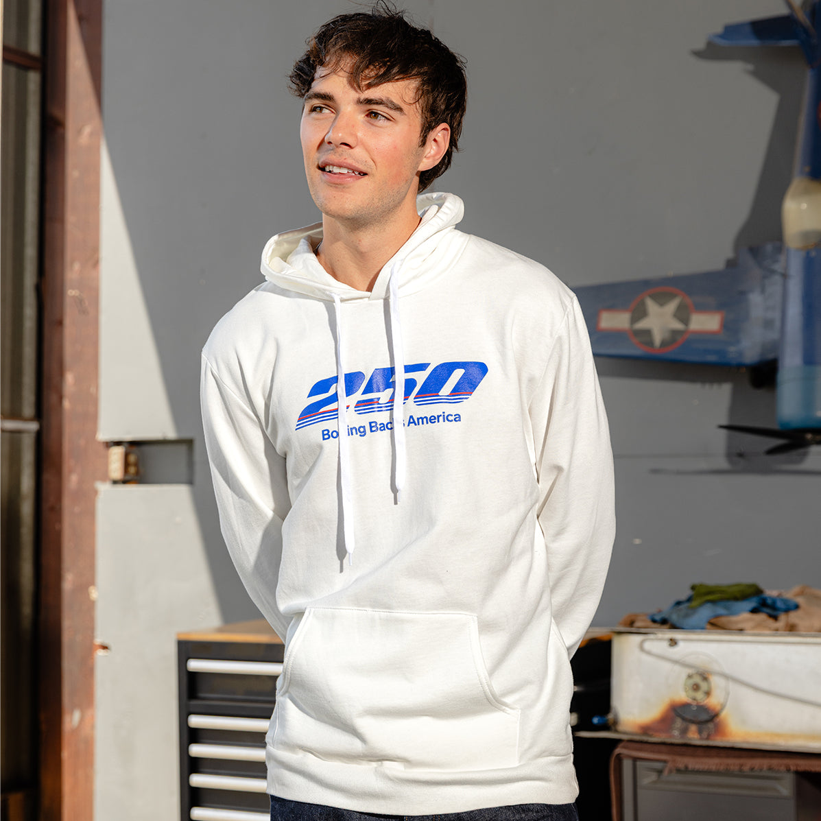 A young man stands indoors, smiling slightly, wearing a BOEING America 250 Unisex Hoodie with blue text. Sunlight streams in from the left, illuminating a workbench, drawers, and a blue model airplane mounted on the wall behind him.