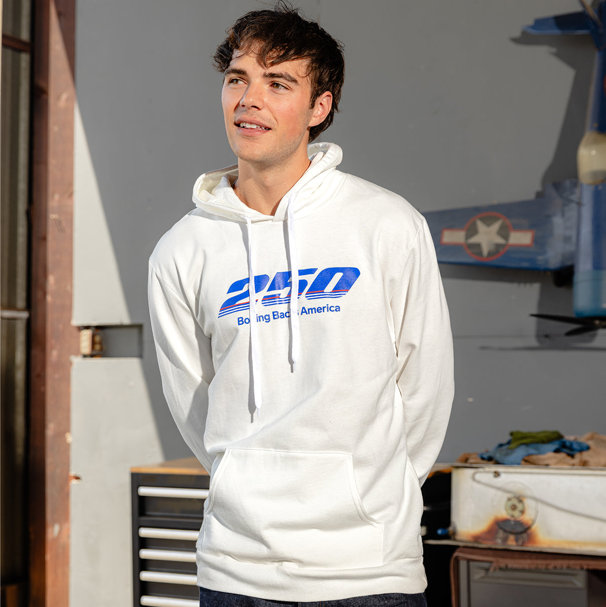 A young man stands indoors, smiling slightly, wearing a BOEING America 250 Unisex Hoodie with blue text. Sunlight streams in from the left, illuminating a workbench, drawers, and a blue model airplane mounted on the wall behind him.
