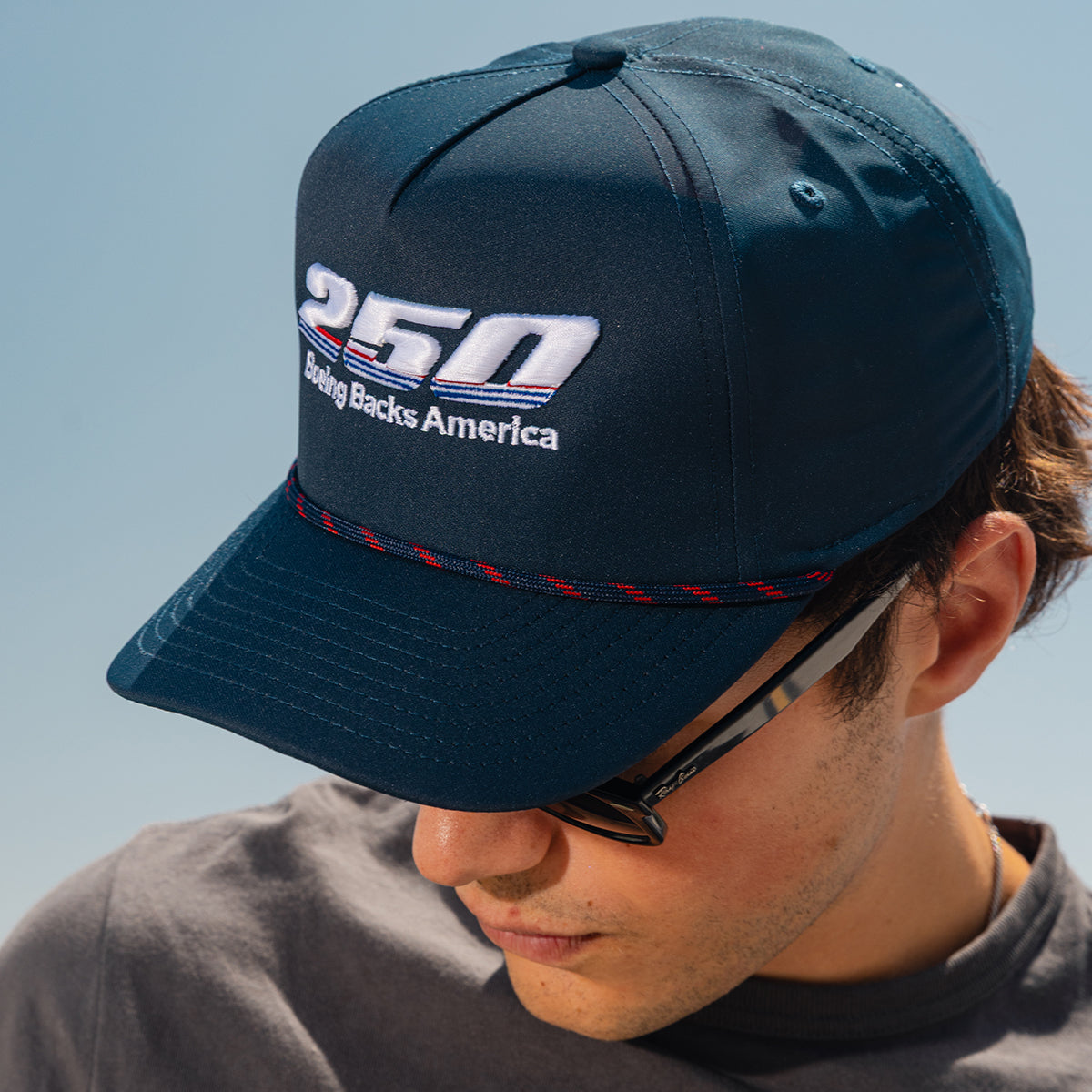 A person wearing sunglasses and a dark blue Boeing America 250 Performance Hat by BOEING, crafted from recycled performance polyester, looks down and enjoys the clear blue sky.