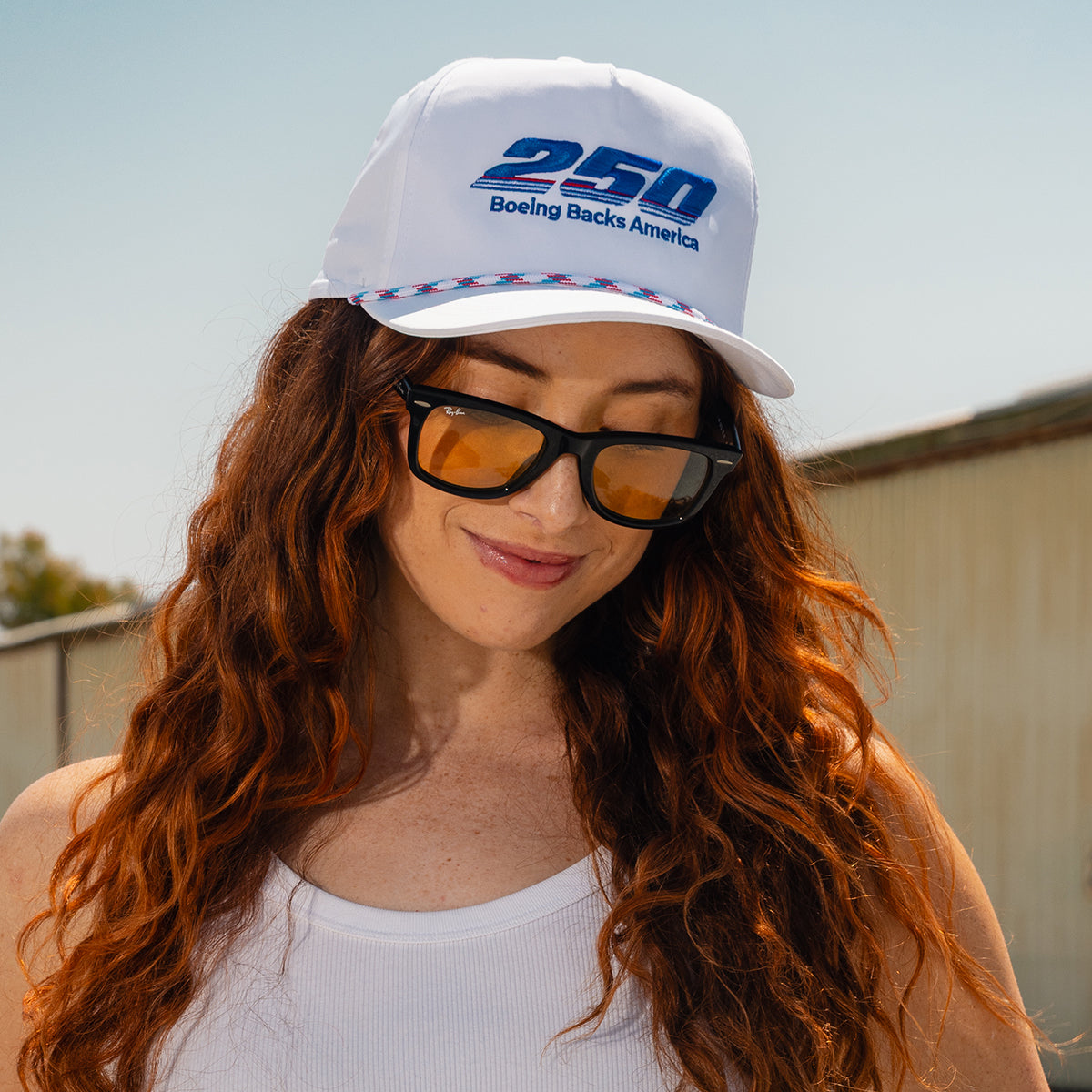 A woman with long red hair wears yellow-tinted sunglasses, a white tank top, and the BOEING America 250 Performance Hat with UPF 50+ protection while standing outdoors on a sunny day.