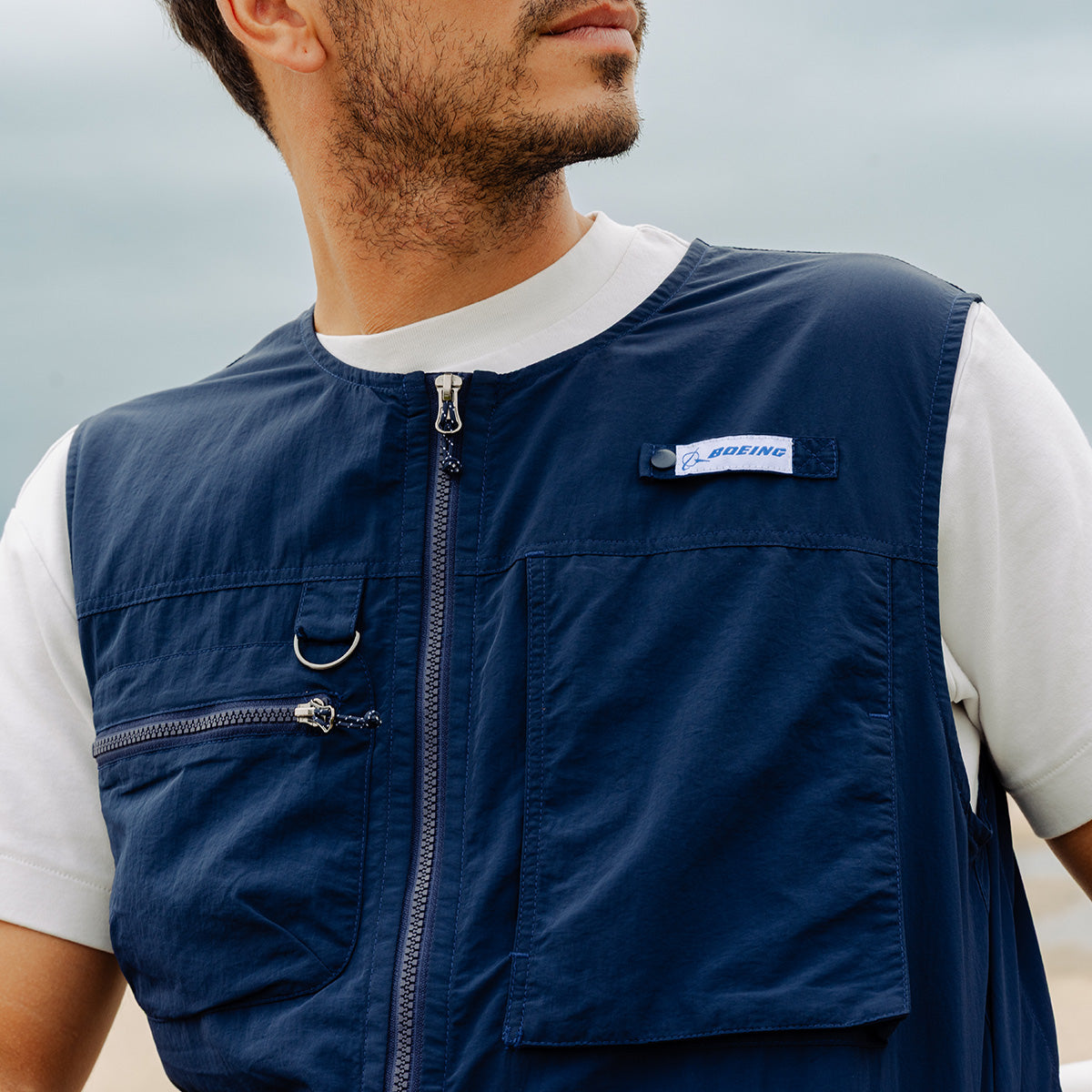 A man stands outdoors under a cloudy sky wearing a white t-shirt and the Boeing Signature Logo Men's Full-Zip Utility Vest by BOEING, a navy blue water-resistant vest with utility pockets and the Boeing logo on the chest.
