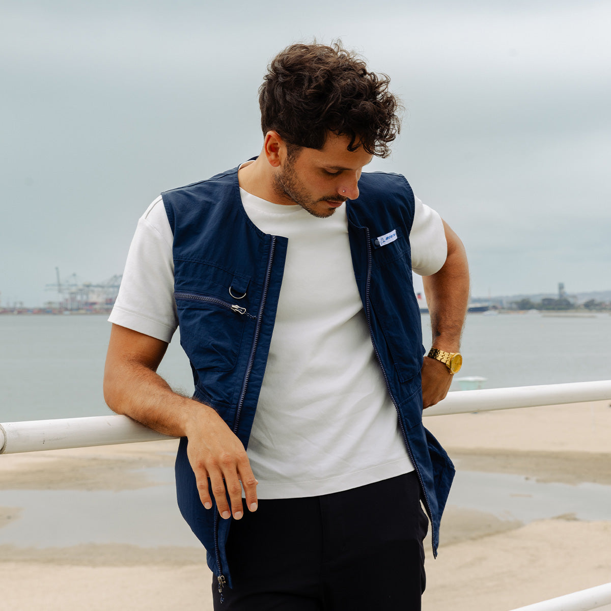 A man in a Boeing Signature Logo Men's Full-Zip Utility Vest by BOEING over a white t-shirt leans against a white railing, looking down, with the beach, water, and an industrial port in the cloudy background.