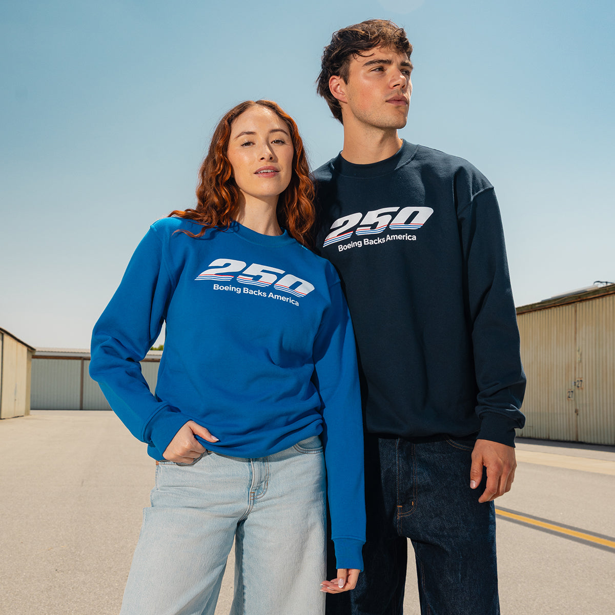 A woman and man pose confidently outdoors in BOEING America 250 Unisex Crewnecks, featuring the “250 Boeing Backs America” design, with industrial buildings and a clear blue sky in the background.