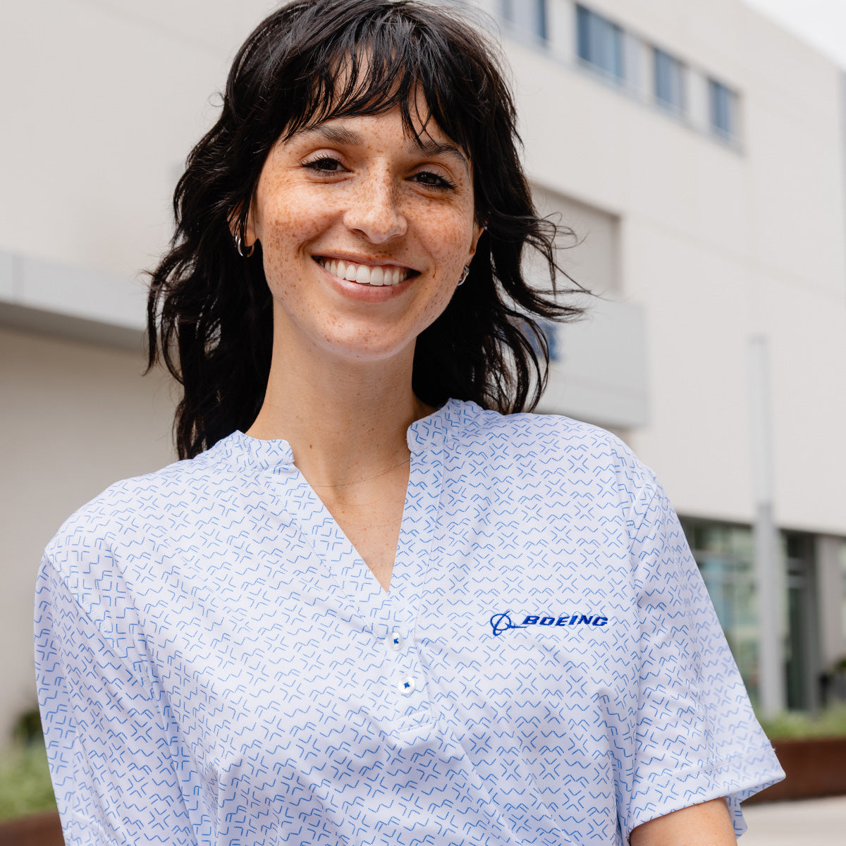 A smiling woman with dark hair and freckles wears the Boeing Women's Signature Logo Pro Crossroads Polo, a white moisture-wicking shirt with a blue BOEING logo, standing outside in front of a modern building.