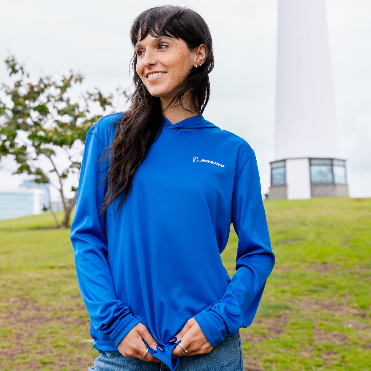 A woman with long dark hair smiles outdoors on grass, wearing a Boeing Signature Logo Women's UV Long Sleeve T-Shirt by Boeing. Trees and a white tower appear in the background.