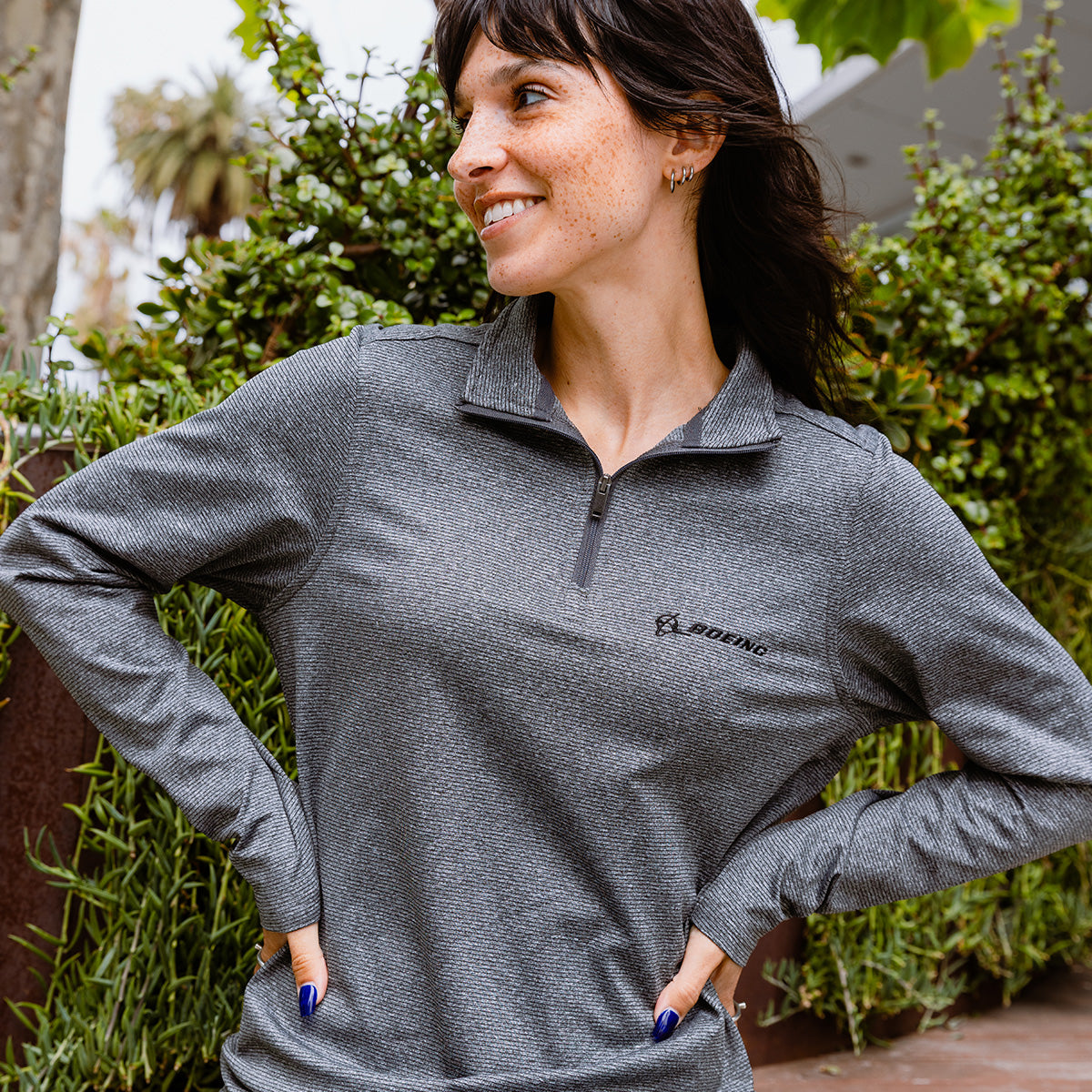 A woman with dark hair and freckles smiles outdoors, wearing a gray Boeing Women's Signature Logo Stripe Quarter-Zip Pullover. Green plants and a tree can be seen in the background.
